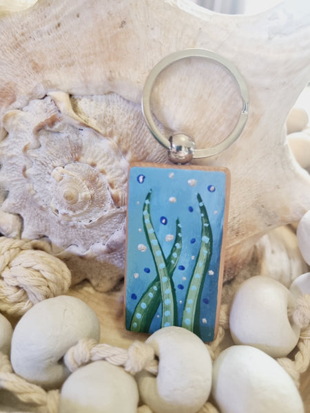 Keychain with a beach-themed design on seashells and rope, hand painted ribbon seaweed on a blue background on a wooden keyring against handmade clay rope garlands by HLSdesigner