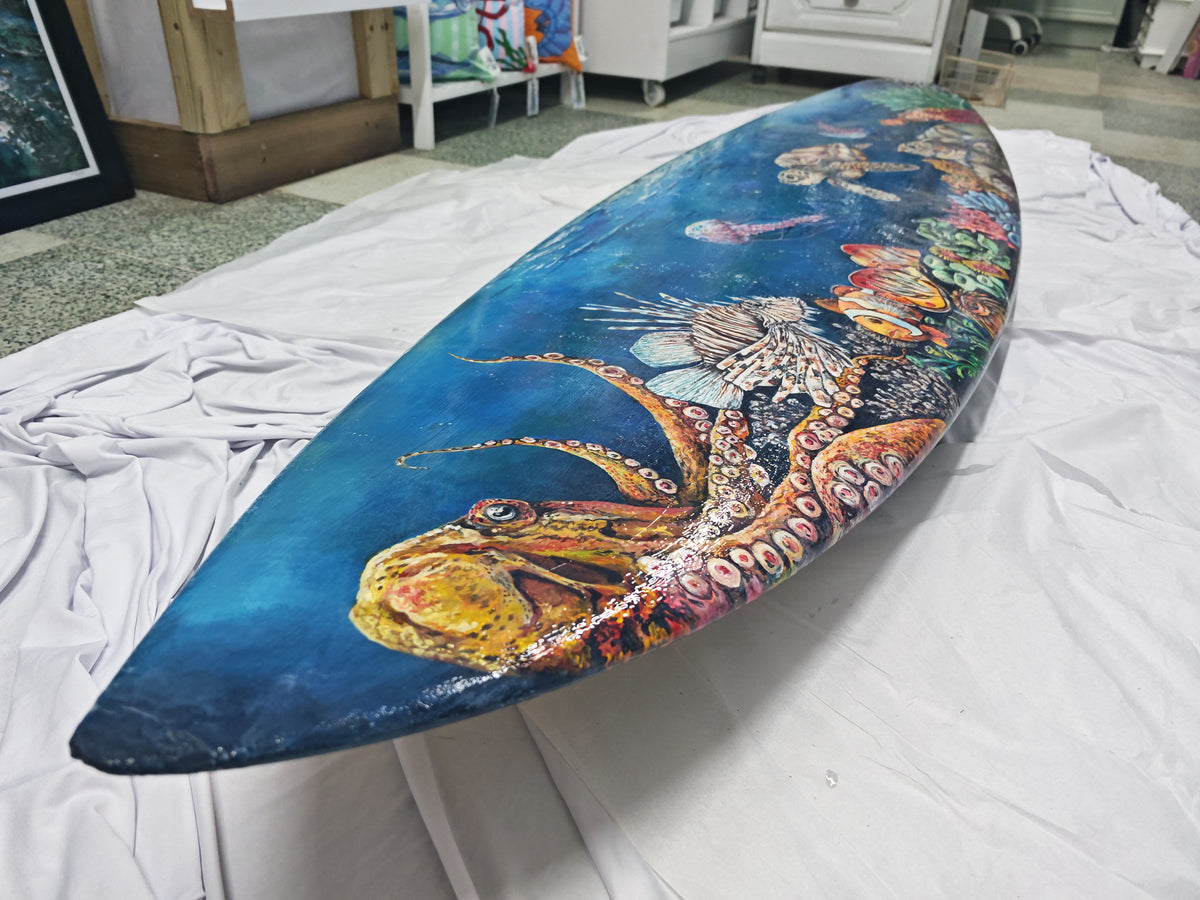 A custom-painted surfboard featuring a detailed illustration of an octopus and sea life on a blue ocean background, displayed on a white sheet in an indoor studio space. Inside the HLS ocean art gallery in Newquay, Original art piece featuring an underwater ocean reef scene only available at HLSdesigner, painted by artist Hannah Singleton, ocean inspired art from Cornwall