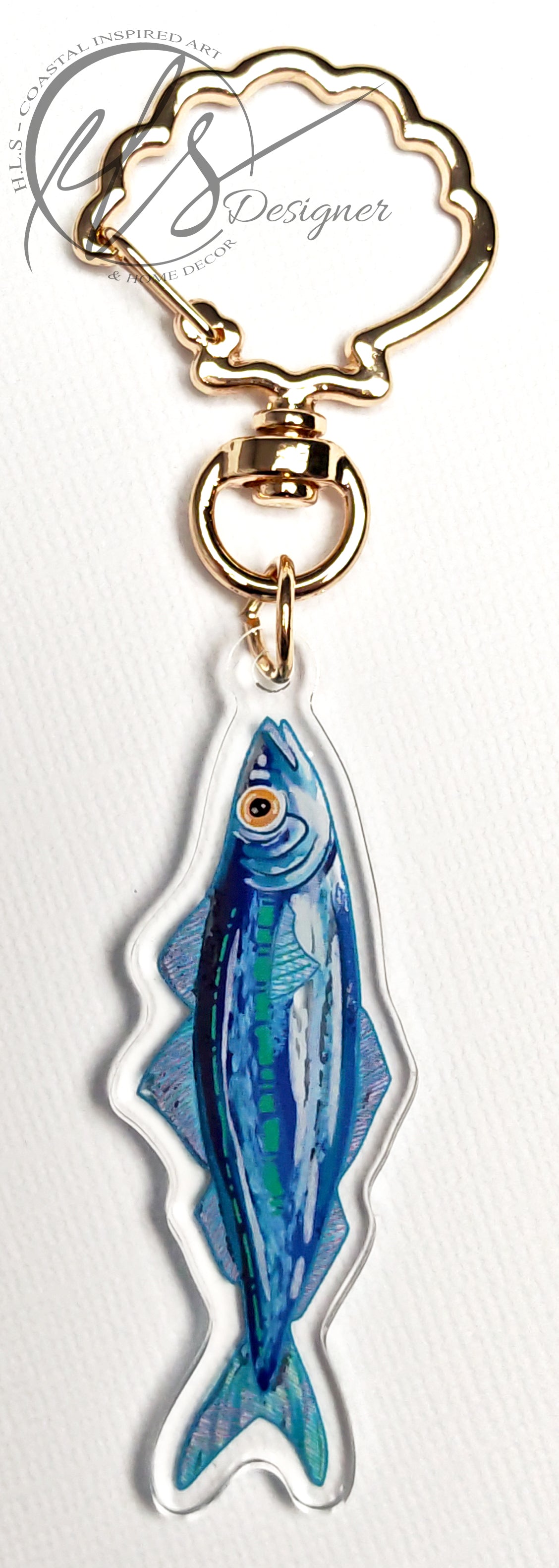watercolour inspired sardine illustration with blues and greens throughout and a bold orange fish eye. Finished in clear acrylic and shaped to the fish with a metal seashell shaped keychain clasp for ocean decoration, image taken on a white background with the HLSdesigner logo in the corner, coastal inspired art and home décor in portrait orientation
