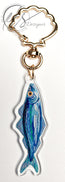 watercolour inspired sardine illustration with blues and greens throughout and a bold orange fish eye. Finished in clear acrylic and shaped to the fish with a metal seashell shaped keychain clasp for ocean decoration, image taken on a white background with the HLSdesigner logo in the corner, coastal inspired art and home décor in portrait orientation