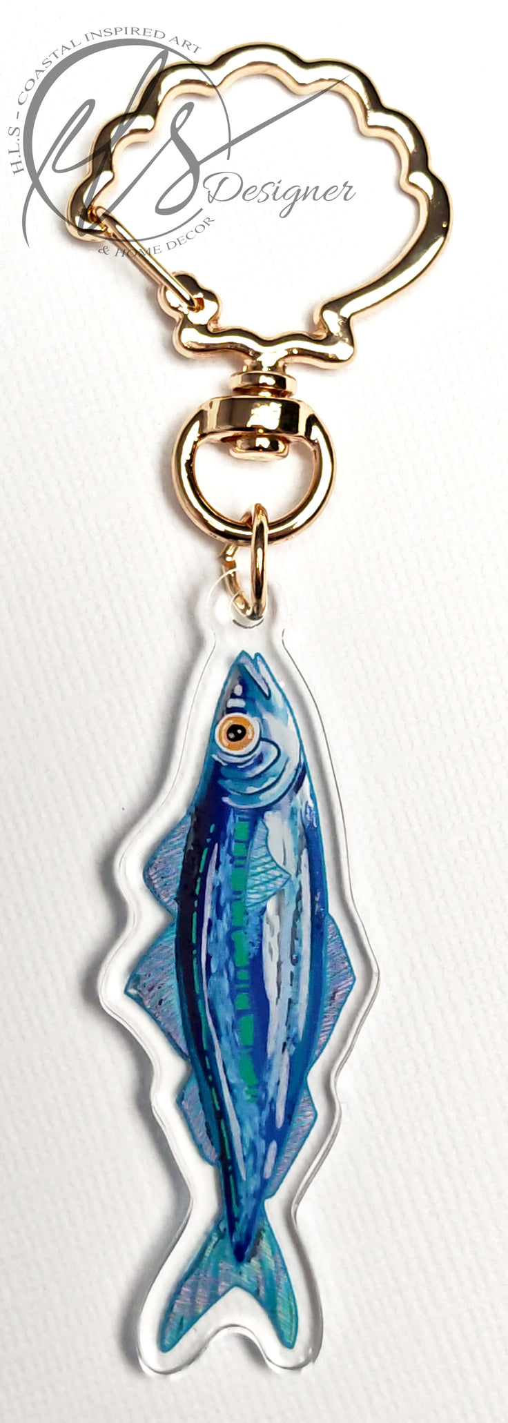 watercolour inspired sardine illustration with blues and greens throughout and a bold orange fish eye. Finished in clear acrylic and shaped to the fish with a metal seashell shaped keychain clasp for ocean decoration, image taken on a white background with the HLSdesigner logo in the corner, coastal inspired art and home décor in portrait orientation