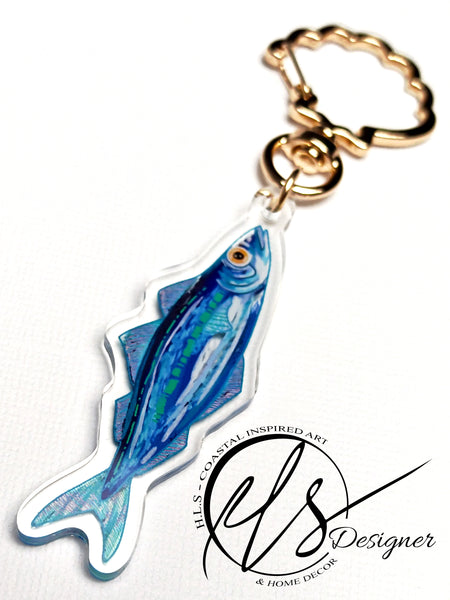 watercolour inspired sardine illustration with blues and greens throughout and a bold orange fish eye. Finished in clear acrylic and shaped to the fish with a metal seashell shaped keychain clasp for ocean decoration, image taken on a white background with the HLSdesigner logo in the corner, coastal inspired art and home décor 