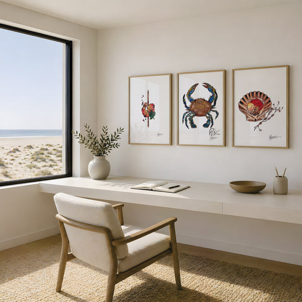 A styled interior room with a white desk, a light wood armchair, and three framed crab and shell artworks hanging on the wall. A large window on the left shows a beach and ocean view. This is Prawn skewer, blue sand crab and scallops - beautifully illustrated works using mixed media, creating a beautiful set of prints to choose from complimenting the entire seafood platter range by HLSdesigner. Hannah Singleton fine artist and illustrator from Cornwall inspired by the ocean and the Cornish coastline 