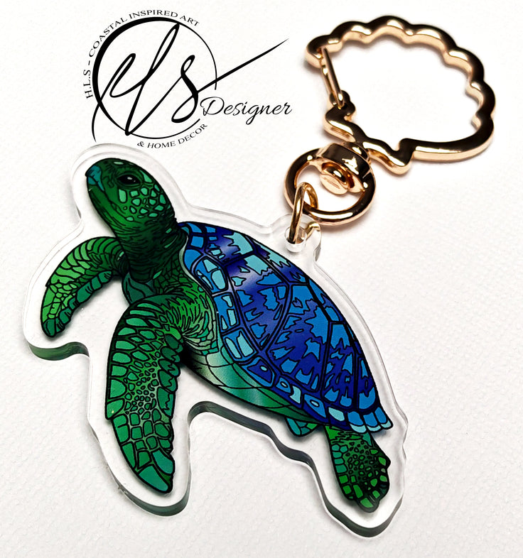 Green and blue sea turtle playful illustration printed into acrylic die cut around and finished with a gold metal seashell shaped keychain clip ring, photo is on a white background with the HLSdesigner logo which reads HLS coastal inspired art and home décor. Designed by HLSdesigner in Cornwall, make your keys, bags and accessories stand out with the ocean collection now