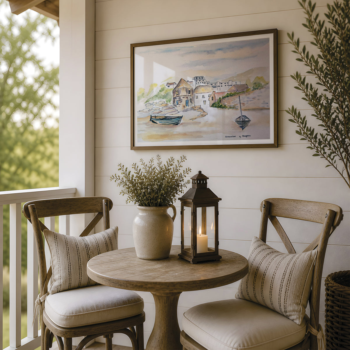 A framed watercolor painting of a coastal village hangs on a white shiplap wall above a small round wooden table on a porch. Two wooden chairs with cream cushions and striped pillows sit beside the table, which holds a ceramic vase with dried stems and a black lantern candle.This picture is a fine art print reproduction of Sennen Cove by Robert Singleton who was known for contemporary watercolour Cornwall townscape paintings. Now available exclusively through HLSdesigner
