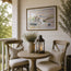A framed watercolor painting of a coastal village hangs on a white shiplap wall above a small round wooden table on a porch. Two wooden chairs with cream cushions and striped pillows sit beside the table, which holds a ceramic vase with dried stems and a black lantern candle.This picture is a fine art print reproduction of Sennen Cove by Robert Singleton who was known for contemporary watercolour Cornwall townscape paintings. Now available exclusively through HLSdesigner