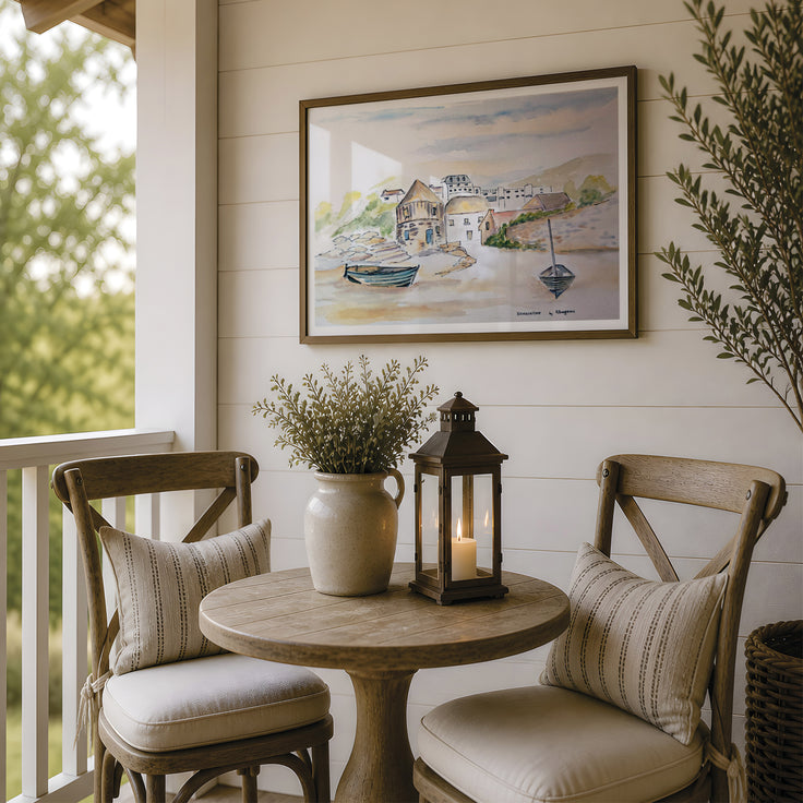 A framed watercolor painting of a coastal village hangs on a white shiplap wall above a small round wooden table on a porch. Two wooden chairs with cream cushions and striped pillows sit beside the table, which holds a ceramic vase with dried stems and a black lantern candle.This picture is a fine art print reproduction of Sennen Cove by Robert Singleton who was known for contemporary watercolour Cornwall townscape paintings. Now available exclusively through HLSdesigner