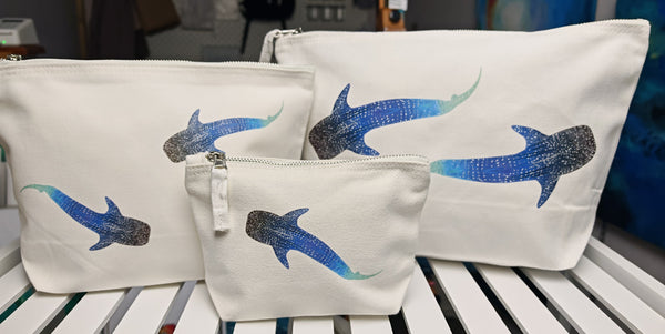 set of three whale shark cosmetic bags, off white cotton canvas accessory bag with metal zip, with ocean themed artwork in the background. whale shark illustration fading from blue to green with white spots pressed onto the bag, the small features one shark and the medium and large bags feature two whale sharks on the bags