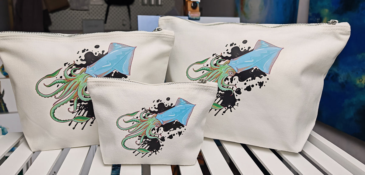 squid ink cosmetic bags, illustration of a squid positioned diagonal with ink splatters behind it and black ink hearts around it, in blue with green and orange tentacles pressed onto an off white cosmetic bag with a silver zip and fabric pull handle.  this is a set of three bags photographed together to compare sizing, in small , medium and large sizes on a white shelf 