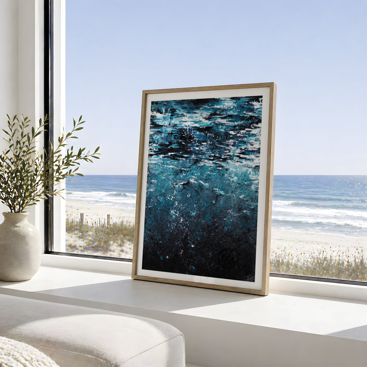 A framed abstract ocean art print with deep blue and black tones, leaning on a white windowsill with a beach and ocean view in the background. Shine from under, an abstract  painting inspired by the serene quietness under the ocean surface bubble rising above you as you ascend to the surface. Contemporary and modern in its statement. Designed by Hannah Singleton at HLSdesigner in Cornwall