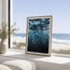 A framed abstract ocean art print with deep blue and black tones, leaning on a white windowsill with a beach and ocean view in the background. Shine from under, an abstract  painting inspired by the serene quietness under the ocean surface bubble rising above you as you ascend to the surface. Contemporary and modern in its statement. Designed by Hannah Singleton at HLSdesigner in Cornwall