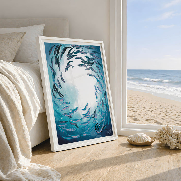 Shoal under surface, a fine art print featuring a shoal of fish swimming around under the ocean surface while the reflections gaze through, with pops of aqua and lilac throughout the colour palette of this print. Designed by HLSdesigner, Hannah Singleton in Cornwall. Ocean inspired fine artist and illustrator. Image features the print in a frame leaning against a bed with a coastal background through a window of the beach house