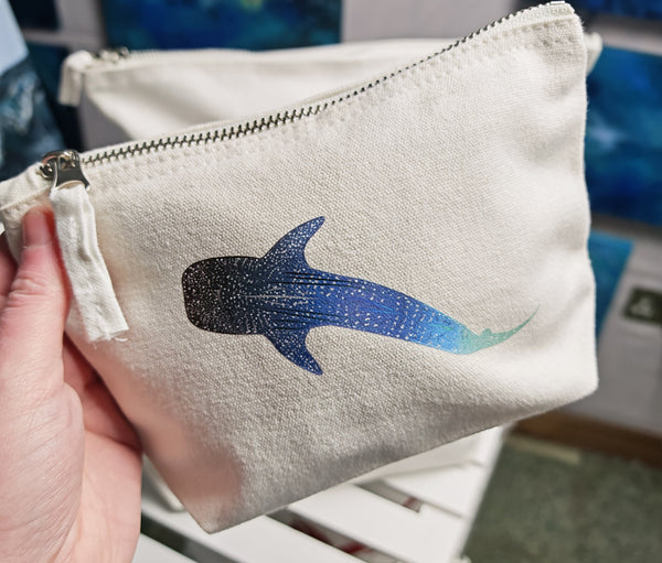small whale shark cosmetic bag, off white cotton canvas accessory bag with metal zip, holding in the photo for size comparison with ocean themed artwork in the background. whale shark illustration fading from blue to green with white spots pressed onto the bag