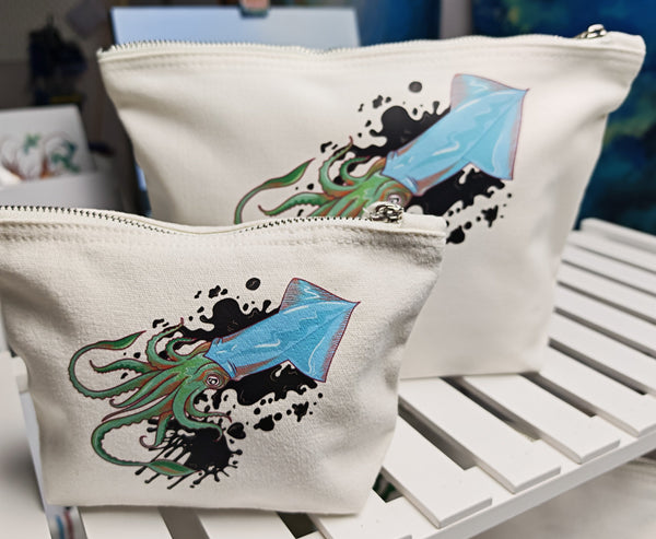 squid ink small and large cosmetic bags, illustration of a squid positioned diagonal with ink splatters behind it and black ink hearts around it, in blue with green and orange tentacles pressed onto an off white cosmetic bag with a silver zip and fabric pull handle.  