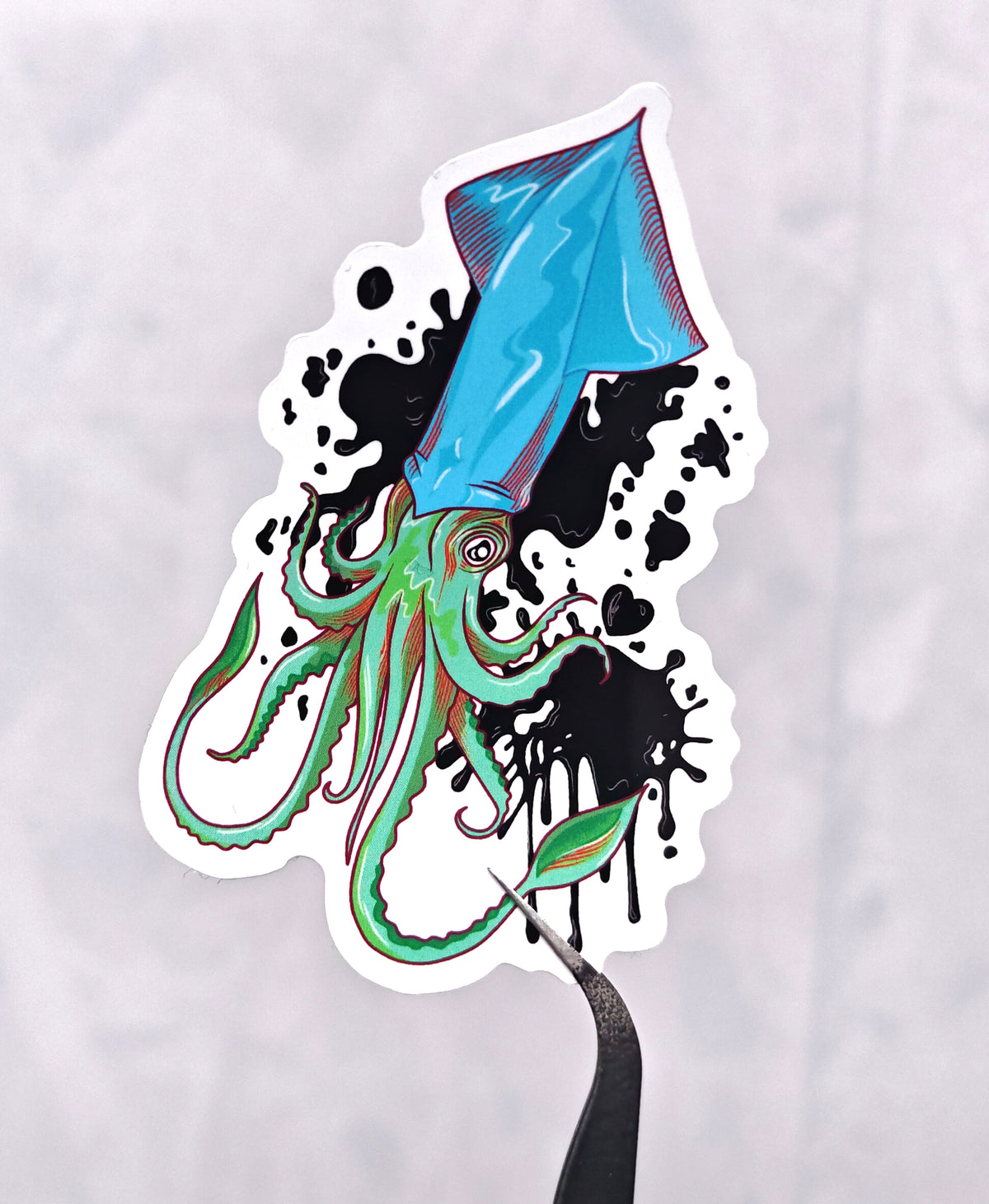 blue and green squid with black ink and black hearts in an ink form surrounding it, illustrated and formed into a die cut vinyl sticker, held with tweezers on a white background