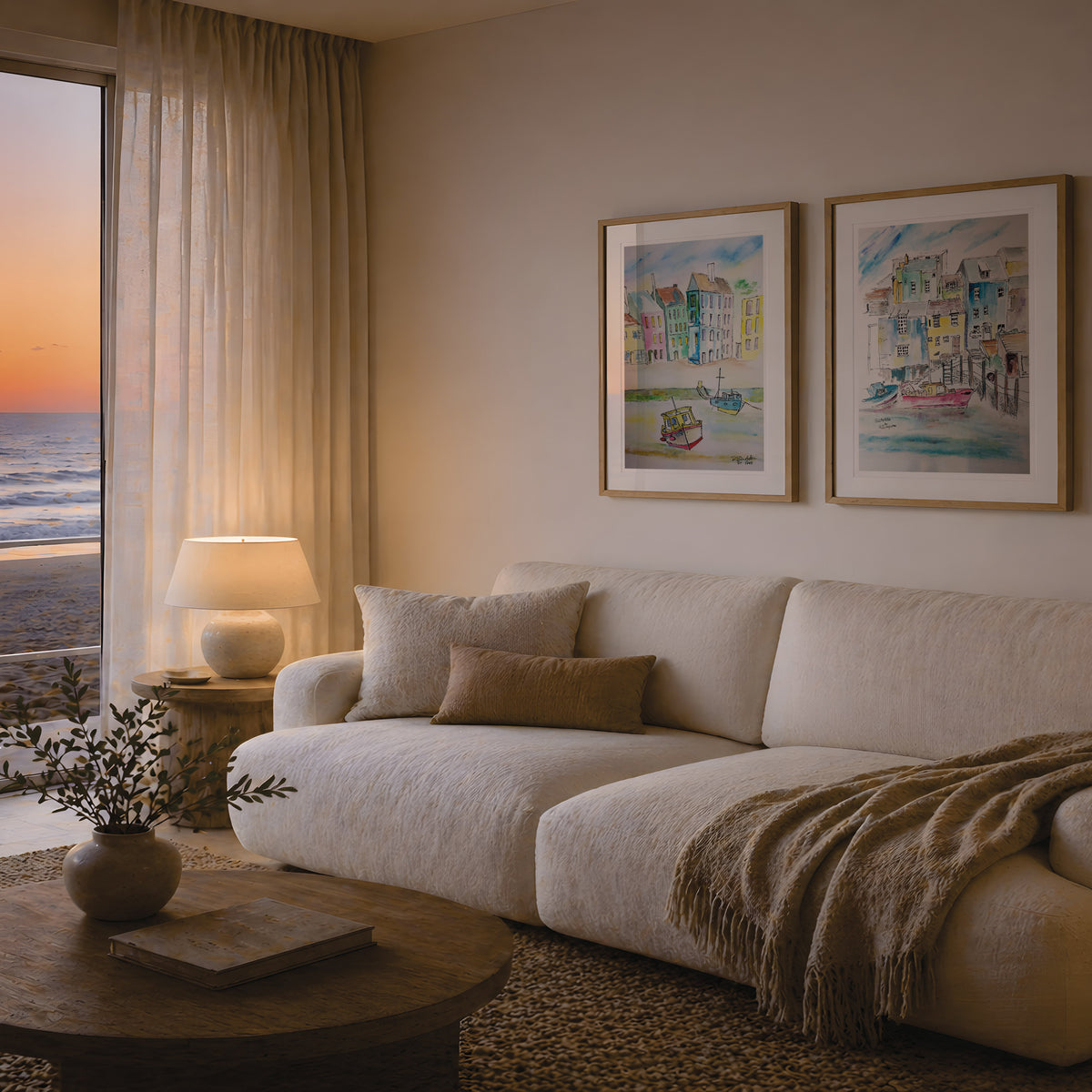 A cozy living room scene with a cream-colored sofa, two framed colorful coastal paintings on the wall, a warm lamp, and a large window showing an ocean sunset. The image appears to be a styled interior scene rather than a clean isolated product shot Displaying fine art prints reproduced by HLSdesigner in memory of contemporary artist Robert Singleton -Creating colourful watercolour studies of popular Cornish towns and coastal scenes. St.IVES and Polperro are featured in this living area