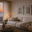 A cozy living room scene with a cream-colored sofa, two framed colorful coastal paintings on the wall, a warm lamp, and a large window showing an ocean sunset. The image appears to be a styled interior scene rather than a clean isolated product shot Displaying fine art prints reproduced by HLSdesigner in memory of contemporary artist Robert Singleton -Creating colourful watercolour studies of popular Cornish towns and coastal scenes. St.IVES and Polperro are featured in this living area