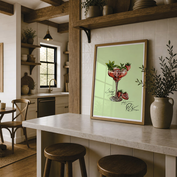 A framed art print featuring a strawberry daiquiri illustration on a light green background, styled on a kitchen island in a rustic kitchen setting. fine art print designed by Hannah Singleton at HLSdesigner in Cornwall. inspired by coastal, ocean and foodie fine art and illustration. Using vivid red shades in a blending mode to create a realistic yet stylised portrayal of a gorgeous summer cocktail