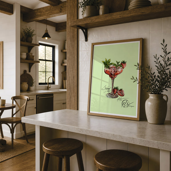A framed art print featuring a strawberry daiquiri illustration on a light green background, styled on a kitchen island in a rustic kitchen setting. fine art print designed by Hannah Singleton at HLSdesigner in Cornwall. inspired by coastal, ocean and foodie fine art and illustration. Using vivid red shades in a blending mode to create a realistic yet stylised portrayal of a gorgeous summer cocktail