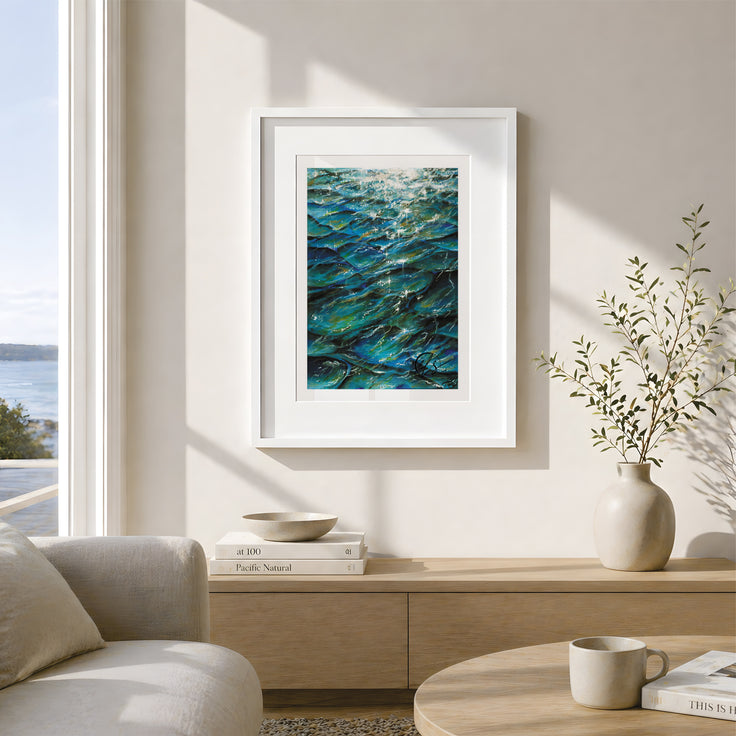 Tranquil hues - a fine art print reproduction of an original painting by HLSdesigner - Hannah Singleton fine artist and illustrator inspired by the ocean and the unique viewpoints of reflections bouncing off the sea surface and underwater. Featuring bold indigo hues and turquoise with green algae blooms throughout, white light reflect off the peaks as the waves slowly move by. Image shows positioned framed on a wall with a coastal theme throughout and an ocean view in the background